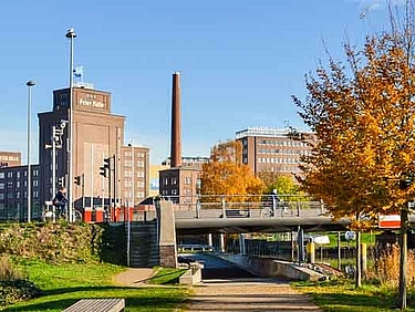 Begrünter Wanderweg am Wasser entlang der Krückau unter einer Brücke und im Hintergrund ist die Stadt Elmshorn zu sehen